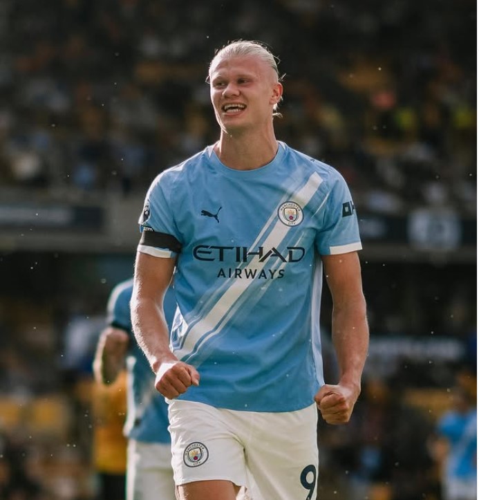 Erling Haaland celebrating after scoring, arms outstretched with a determined expression, teammates approaching to congratulate him, Etihad Stadium crowd visible in the background, energetic and triumphant mood